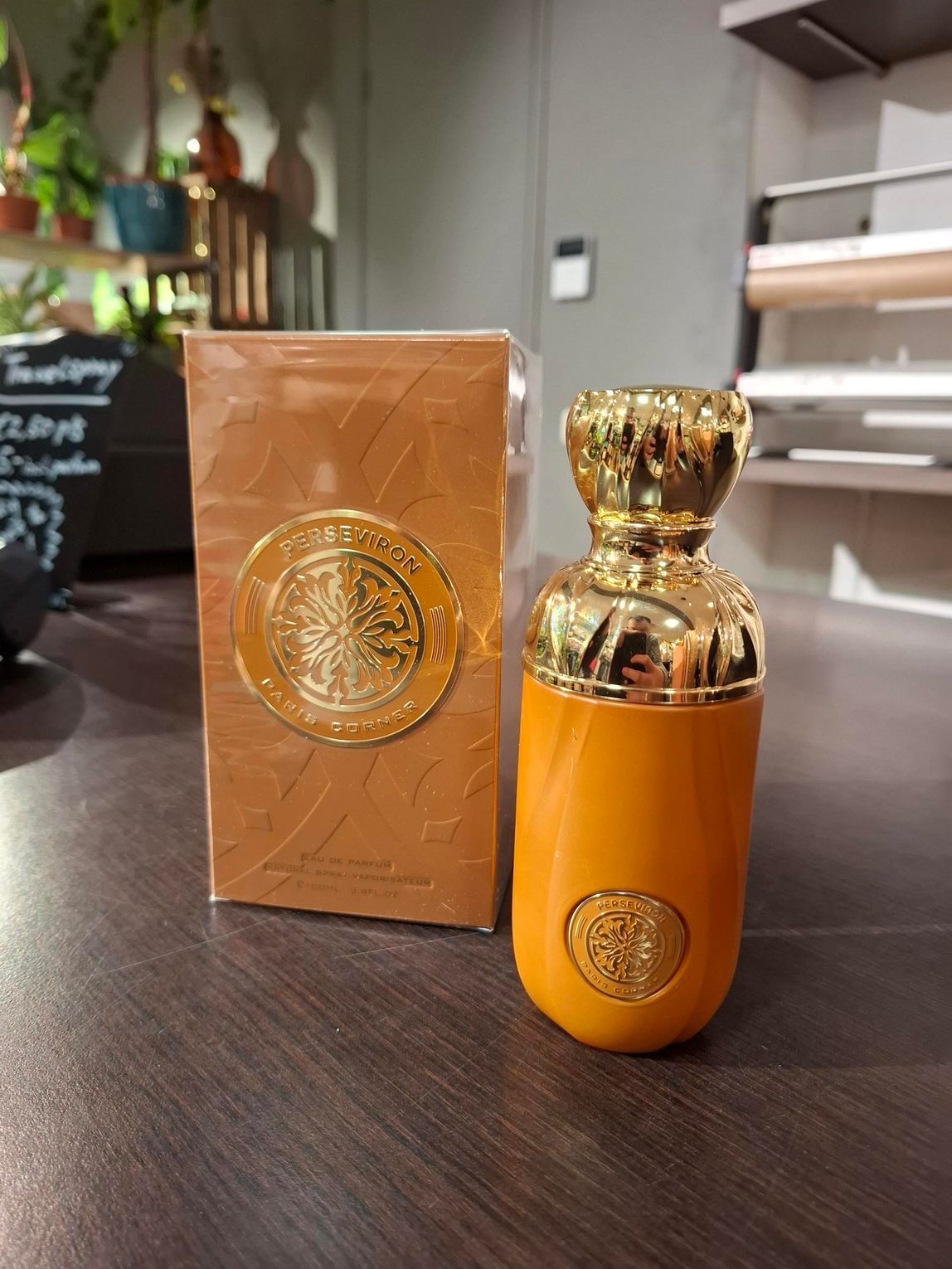 Een flesje oranje parfum met een gouden dop, naast het bijbehorende doosje, staat op een donkerhouten ondergrond.