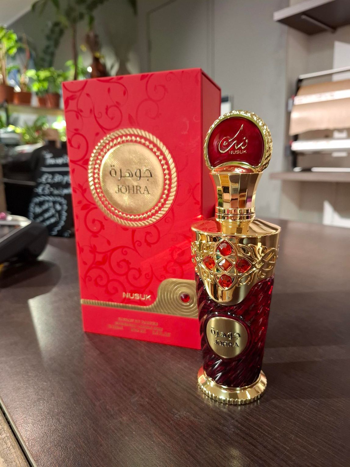 Een rood-gouden parfumflesje van Nabeel Johra staat naast de bijpassende rode decoratieve doos op een houten ondergrond.
