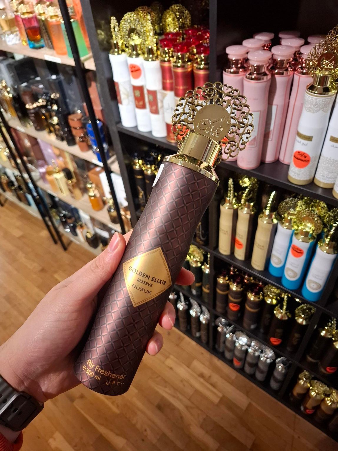 In een winkel met schappen vol parfums houdt een hand een bruine, gestructureerde parfumfles vast met een decoratieve gouden dop.