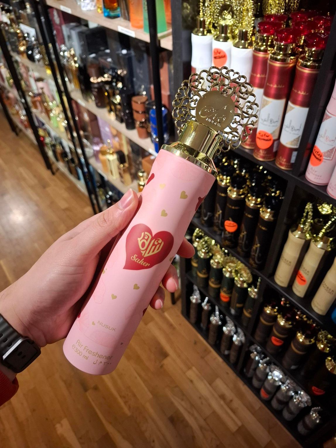 In een winkel houdt een hand een roze parfumverstuiver met hartjespatroon en een sierlijke gouden dop vast.