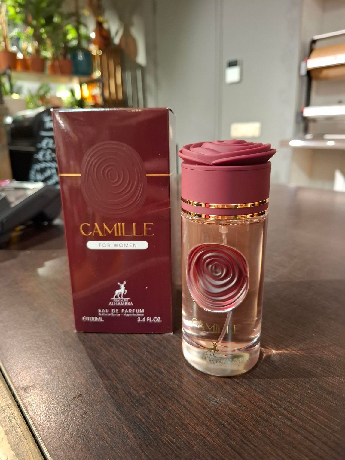 Een flesje Camille Eau de Parfum en de bijbehorende bordeauxrode doos met rozenembleem, staan ​​op een houten ondergrond.