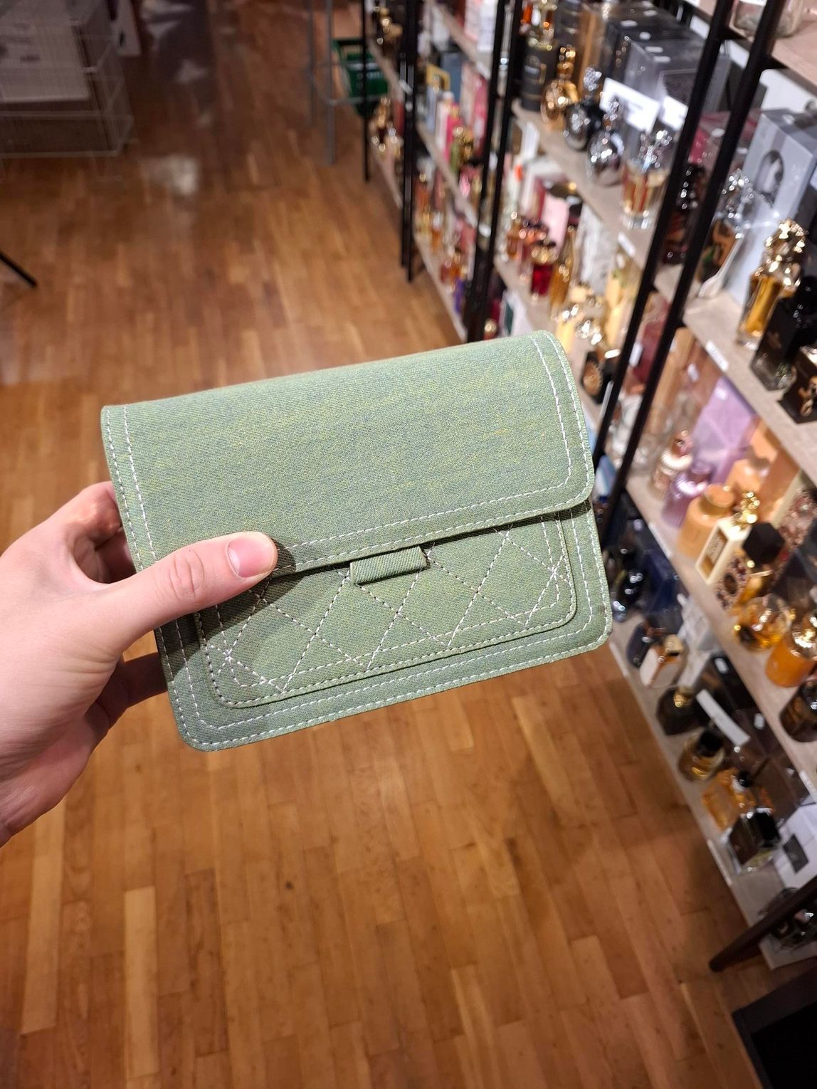 Hand met een kleine groene geweven clutch in een winkelgang met schappen vol producten.
