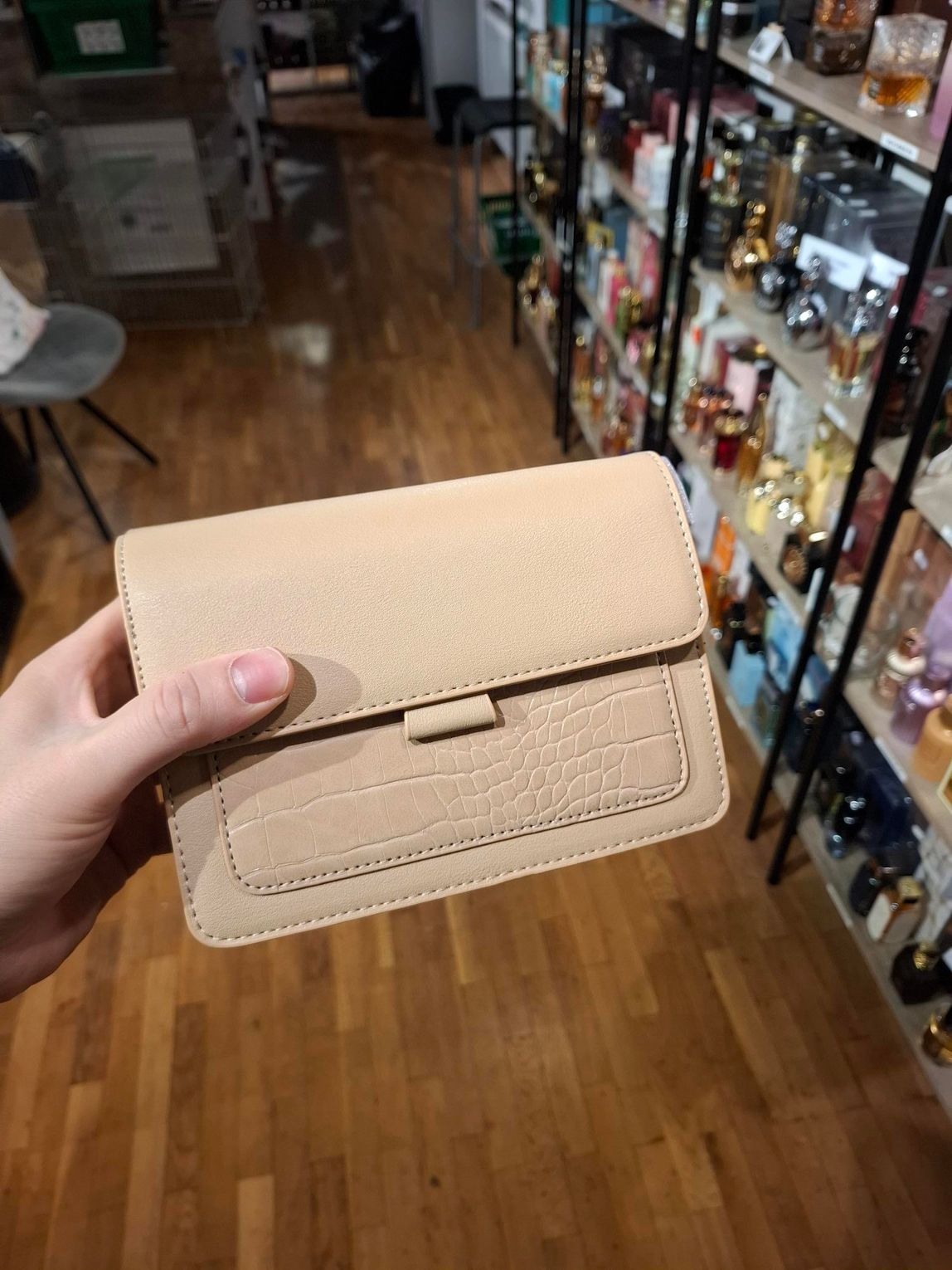 Hand die een beige portemonnee vasthoudt in een winkelgang met schappen vol drank op de achtergrond.