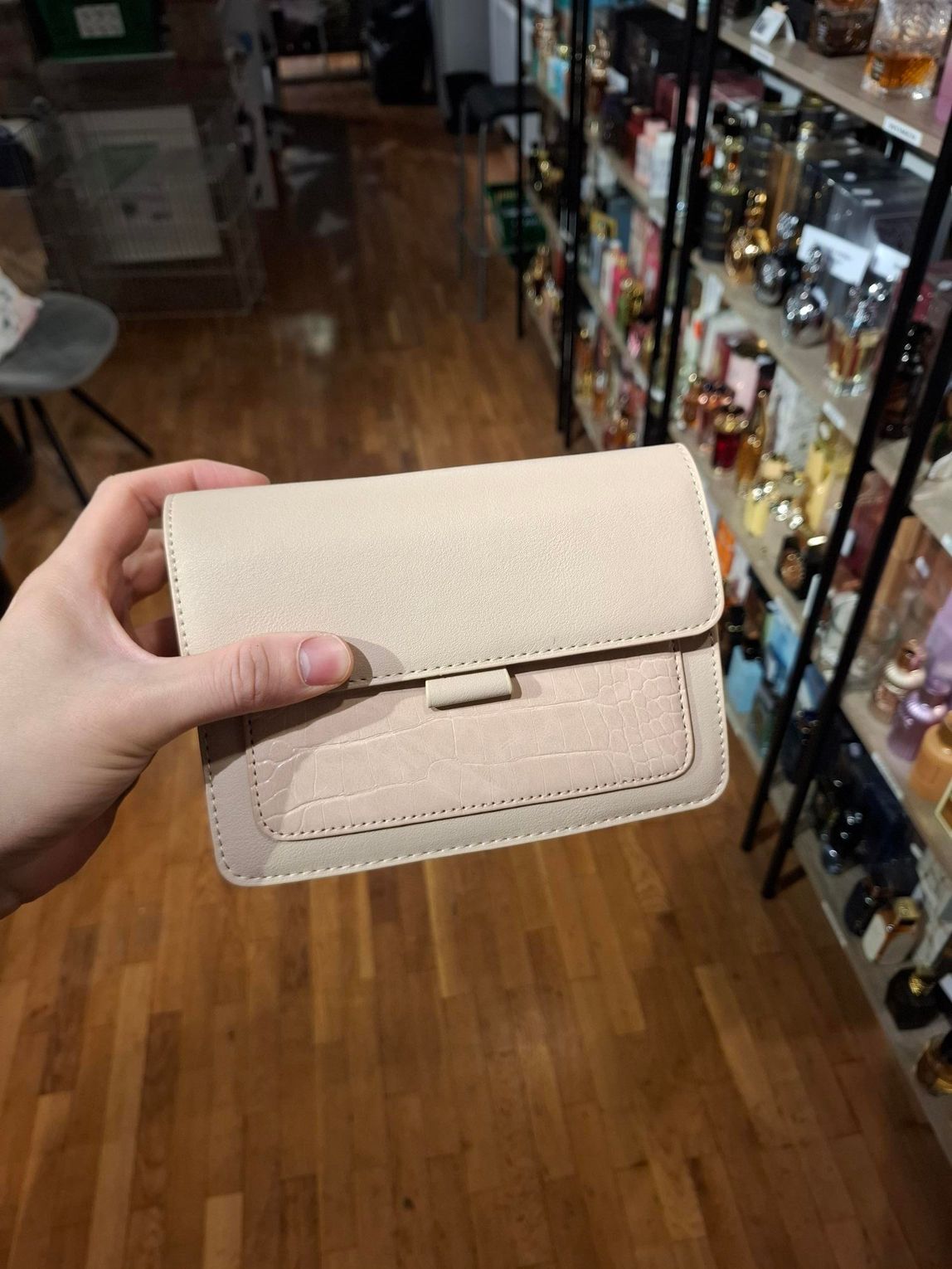 Hand die een beige portemonnee vasthoudt in een winkelgang met schappen vol flessen op de achtergrond.