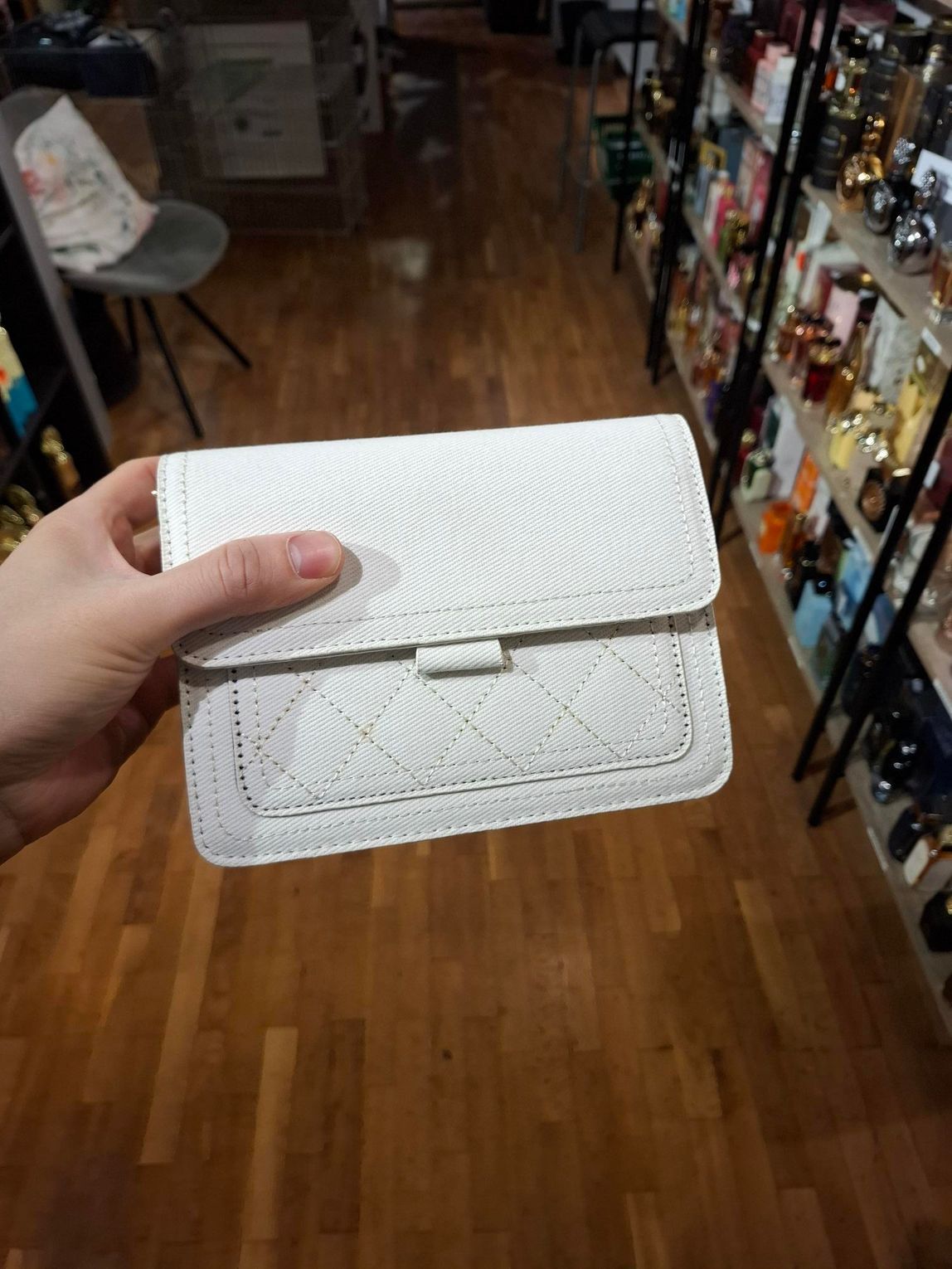 Hand die een witte, gestructureerde clutch vasthoudt in een winkelgang met schappen vol accessoires.