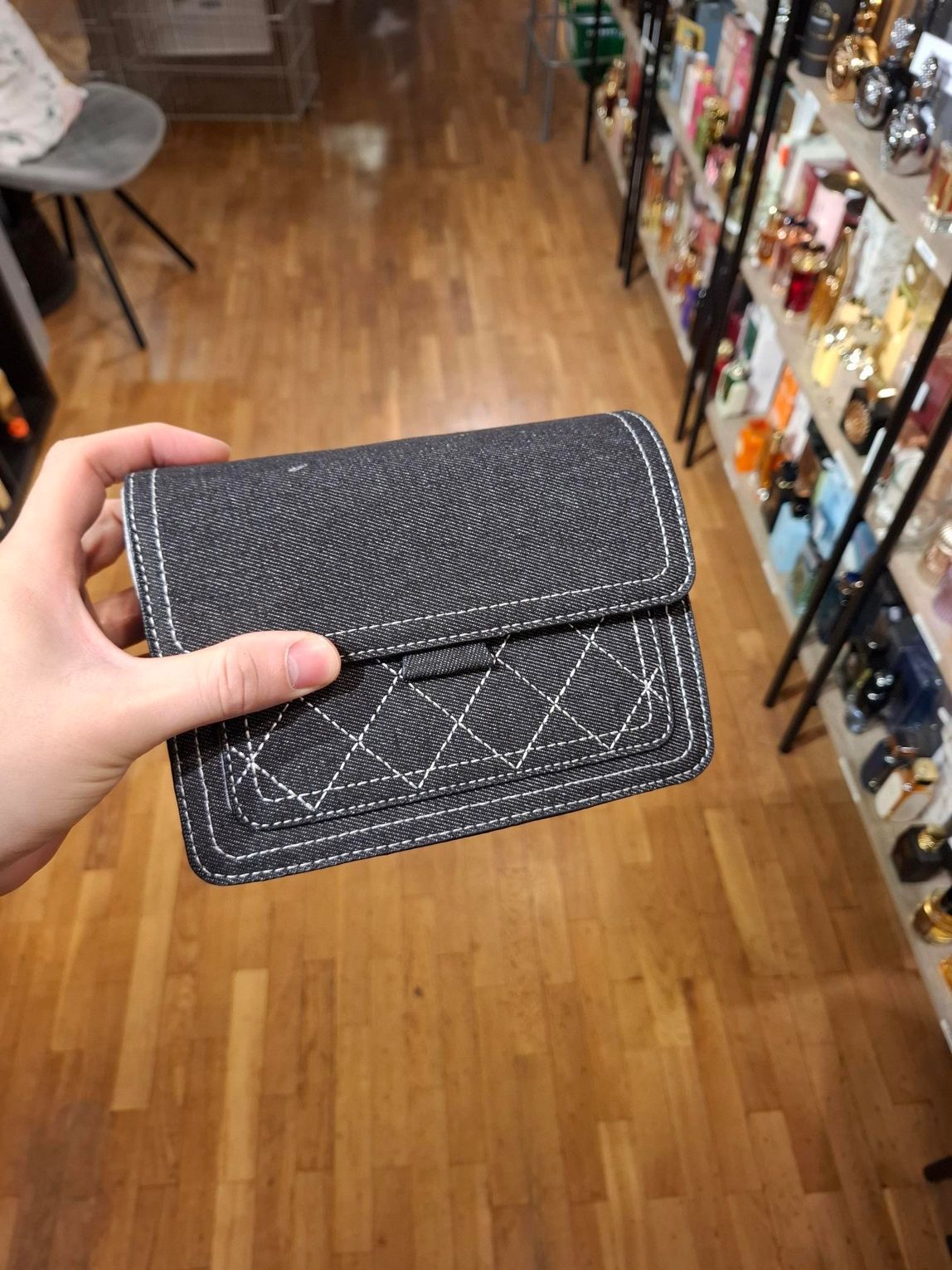 Hand die een zwarte, gewatteerde portemonnee vasthoudt in een winkelgang met een houten vloer en schappen vol goederen.