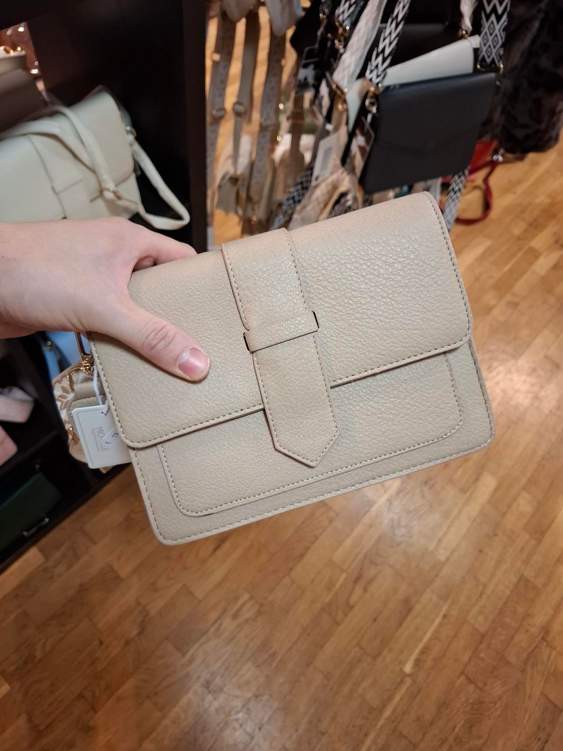Hand die een beige, gestructureerde clutch vasthoudt boven een houten vloer.
