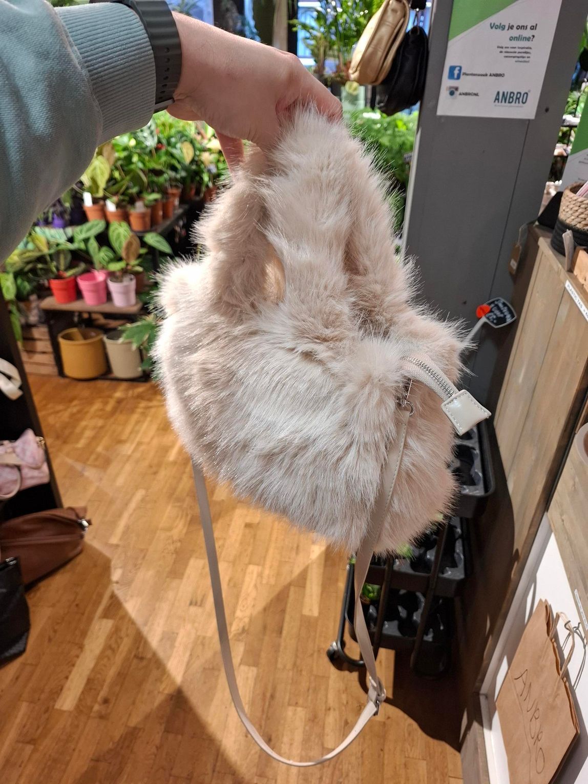 In een winkelomgeving houdt een hand een pluizige, crèmekleurige handtas van imitatiebont met een dun schouderbandje omhoog.