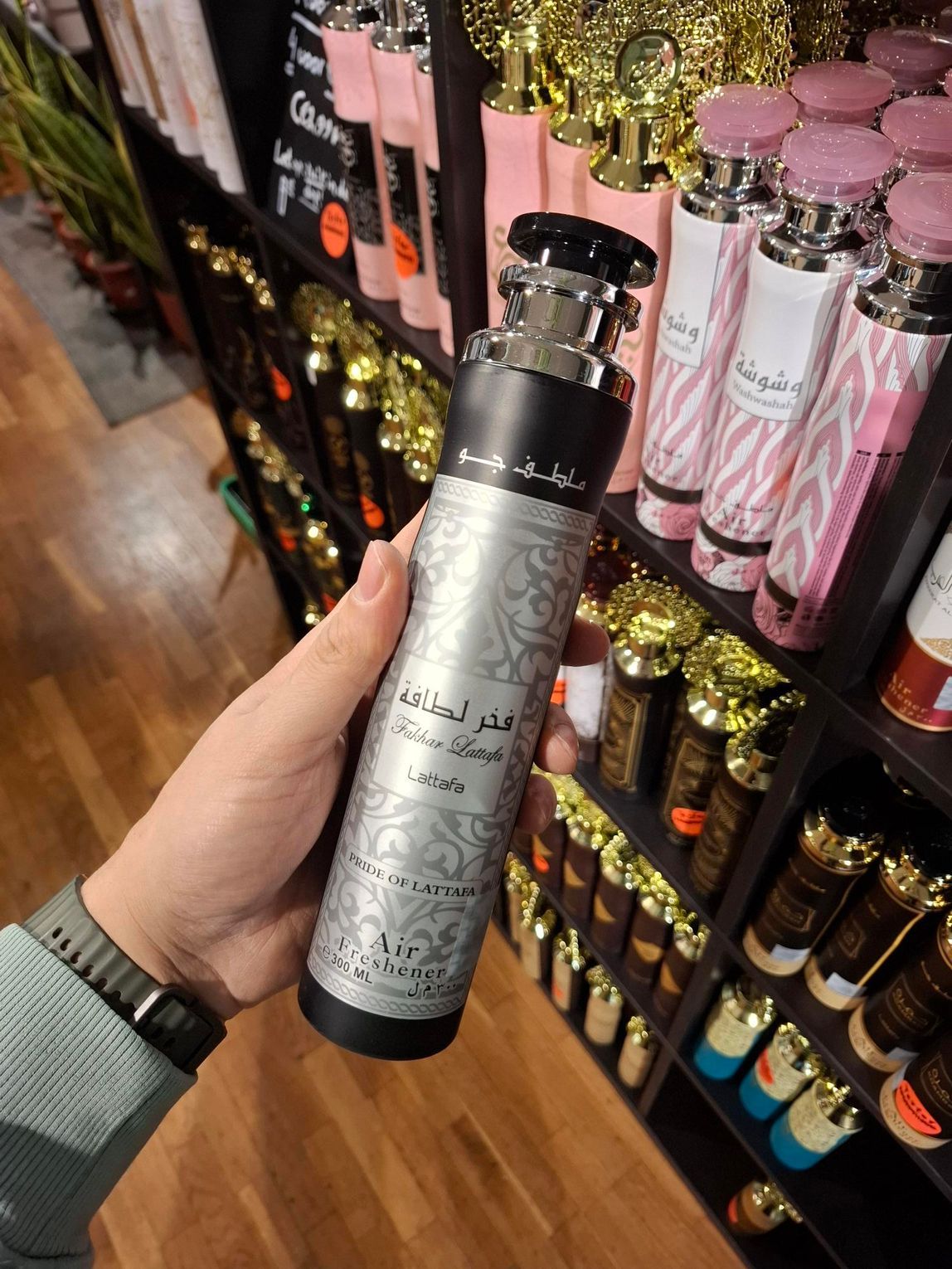 In een winkel met schappen vol producten houdt een hand een hoge, zilverkleurige en zwarte parfumfles met sierlijke patronen vast.