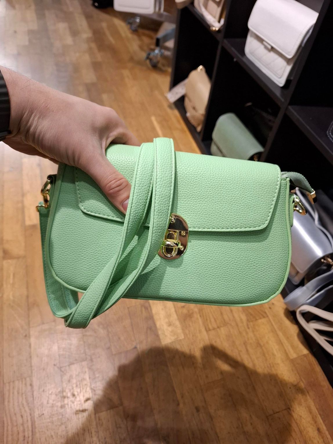 Hand die een mintgroene gewatteerde handtas met gouden sluiting vasthoudt, op de achtergrond van een winkelschap.