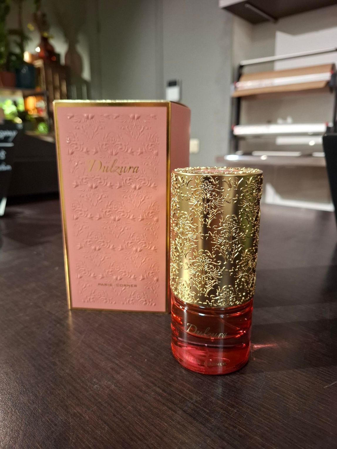 Een roze parfumdoos en een cilindervormige fles met een goudkleurige, gestructureerde dop staan ​​op een donkerhouten ondergrond.