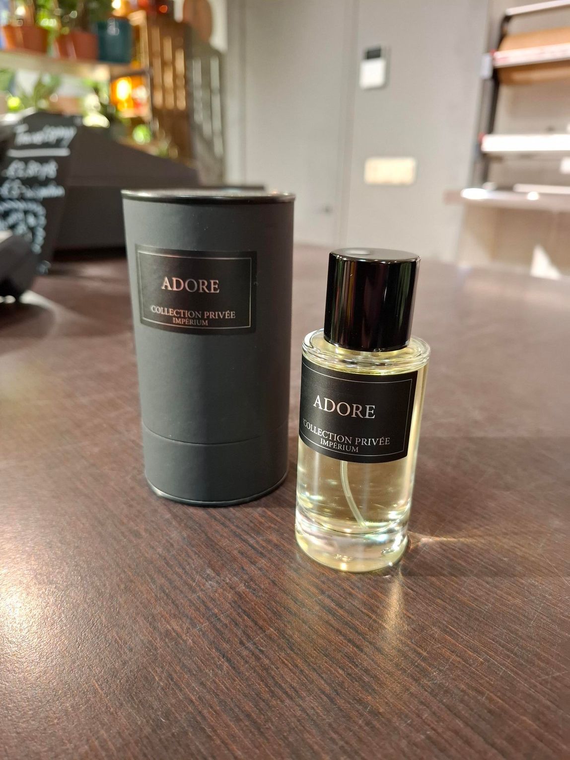 Naast een zwarte, cilindervormige parfumtube staat een bijpassend klein glazen flesje met een zwart etiket waarop ADORE staat.