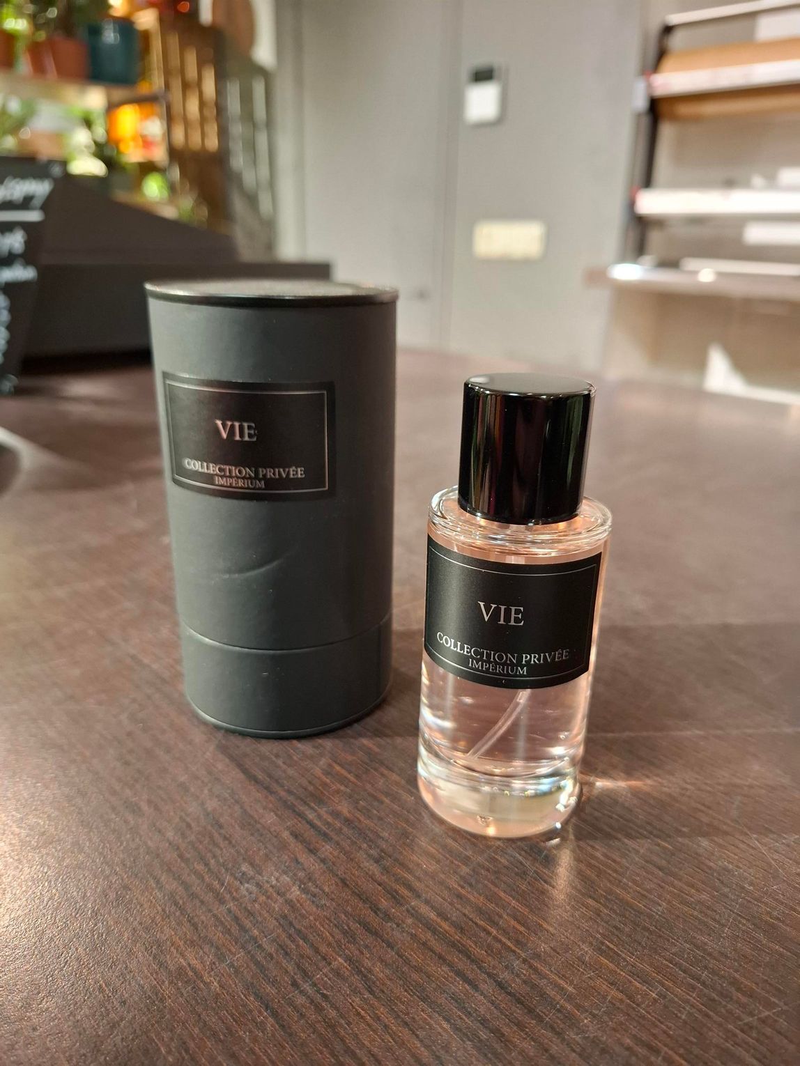 Naast een zwarte, cilindervormige parfumtube staat een bijpassend klein glazen flesje met een zwart etiket waarop ADORE staat.