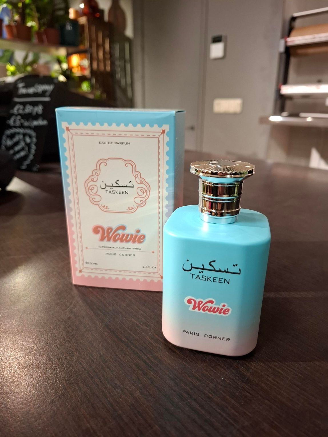 Een flesje Wowie Taskeen-parfum naast de lichtblauwe en roze verpakking, staand op een donkerhouten oppervlak.