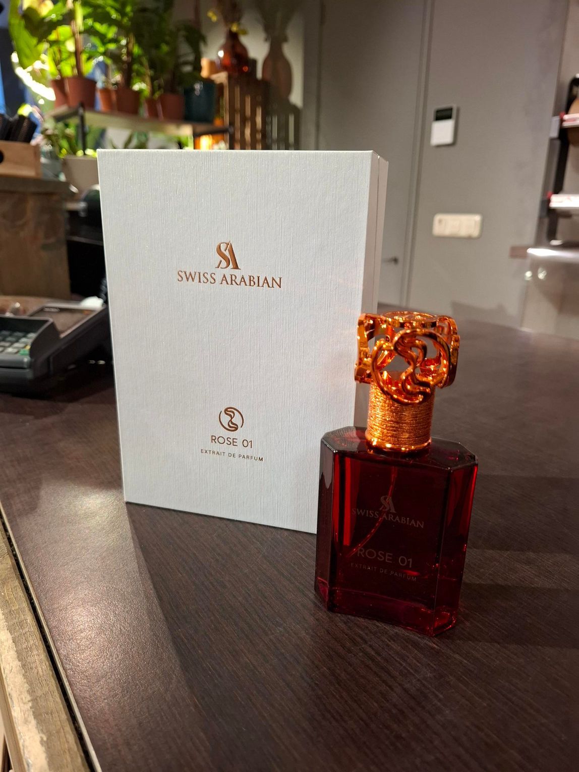 Een flesje Zwitsers Arabisch parfum met een sierlijke koperen dop, naast de witte doos op een houten bureau.