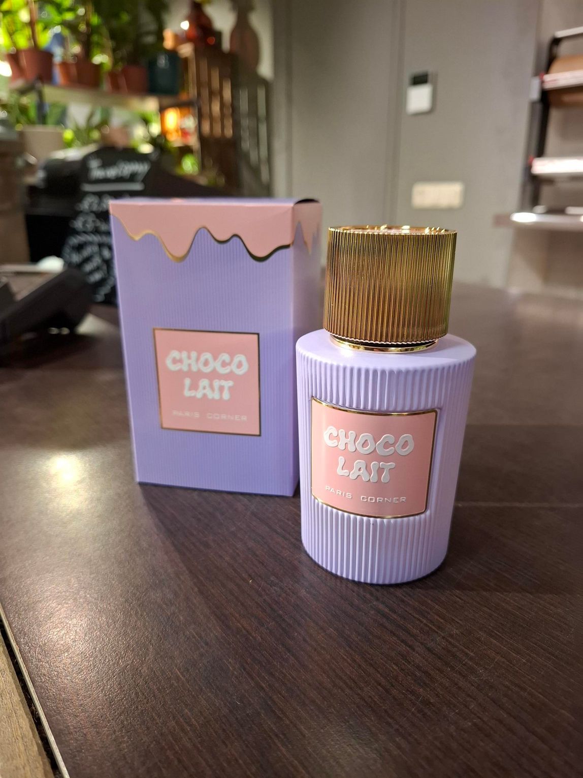 Een flesje Choco Lait-parfum en de bijbehorende doos, beide in pastelpaars met roze etiketten, staan ​​op een houten ondergrond.