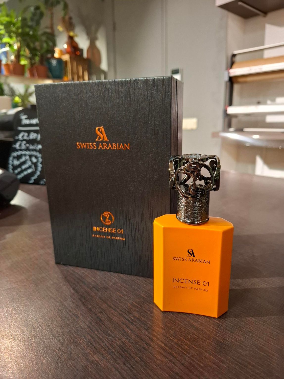 Een zwarte Zwitserse Arabische parfumdoos staat naast een oranje fles met een sierlijke, metalen dop op een donkere ondergrond.