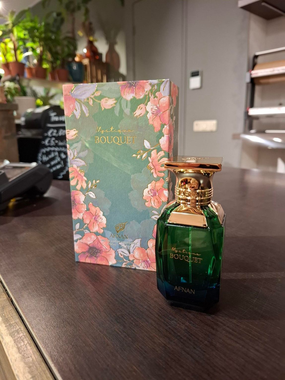 Een groene parfumfles met een gouden dop staat op een houten aanrecht naast de bijpassende doos met bloemenmotief.