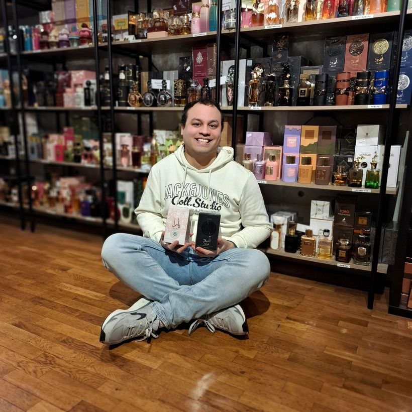 Een persoon zit met gekruiste benen op een houten vloer in een winkel en houdt twee parfumdoosjes vast voor een schap vol flesjes.