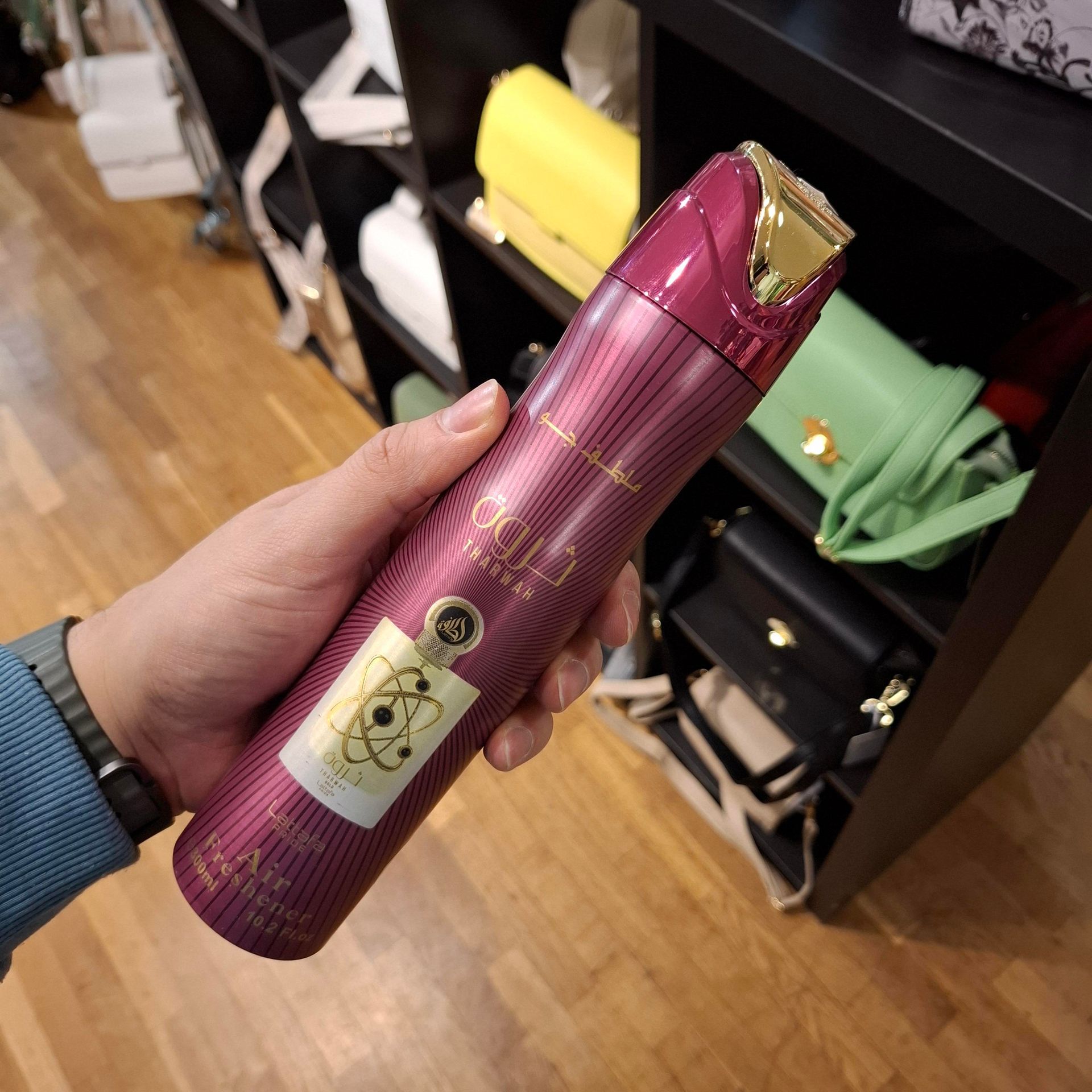 Een hand houdt een magenta spuitbus met luchtverfrisser en gouden accenten vast voor een winkelrek met handtassen.