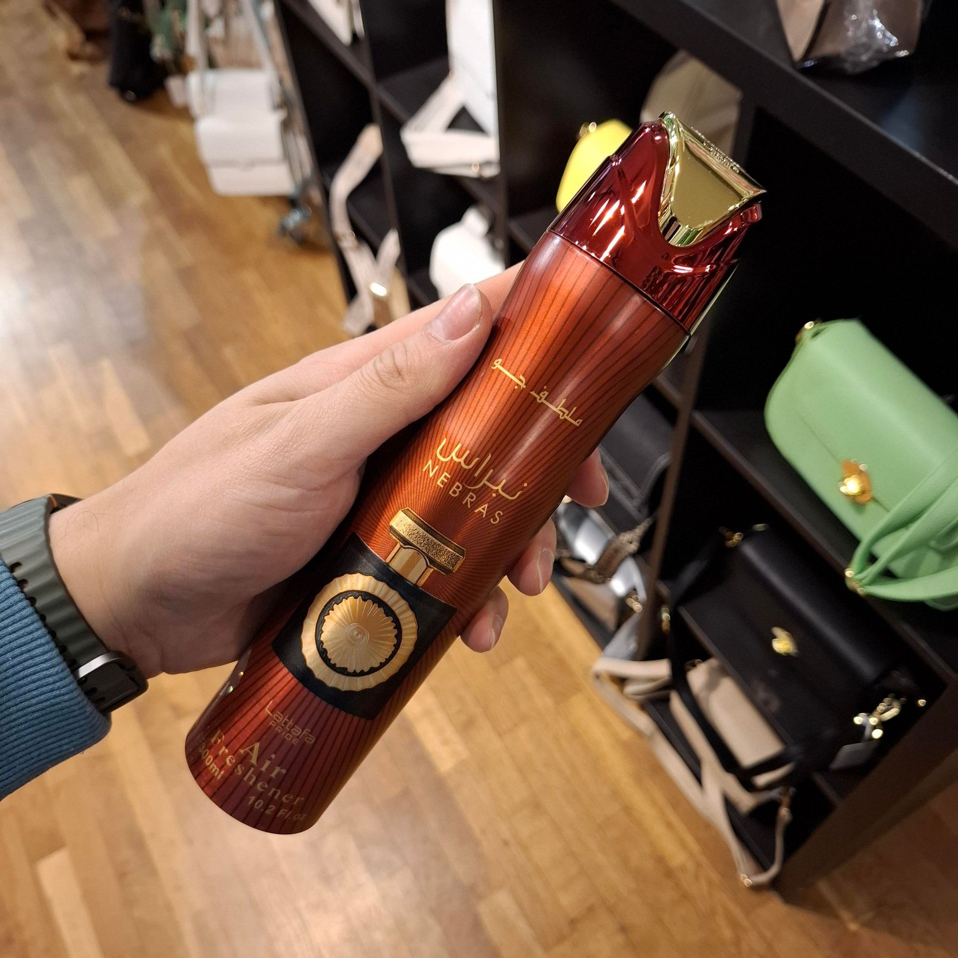 Een hand houdt een hoge, metallic oranje spuitbus met luchtverfrisser vast in een winkelomgeving met schappen op de achtergrond.