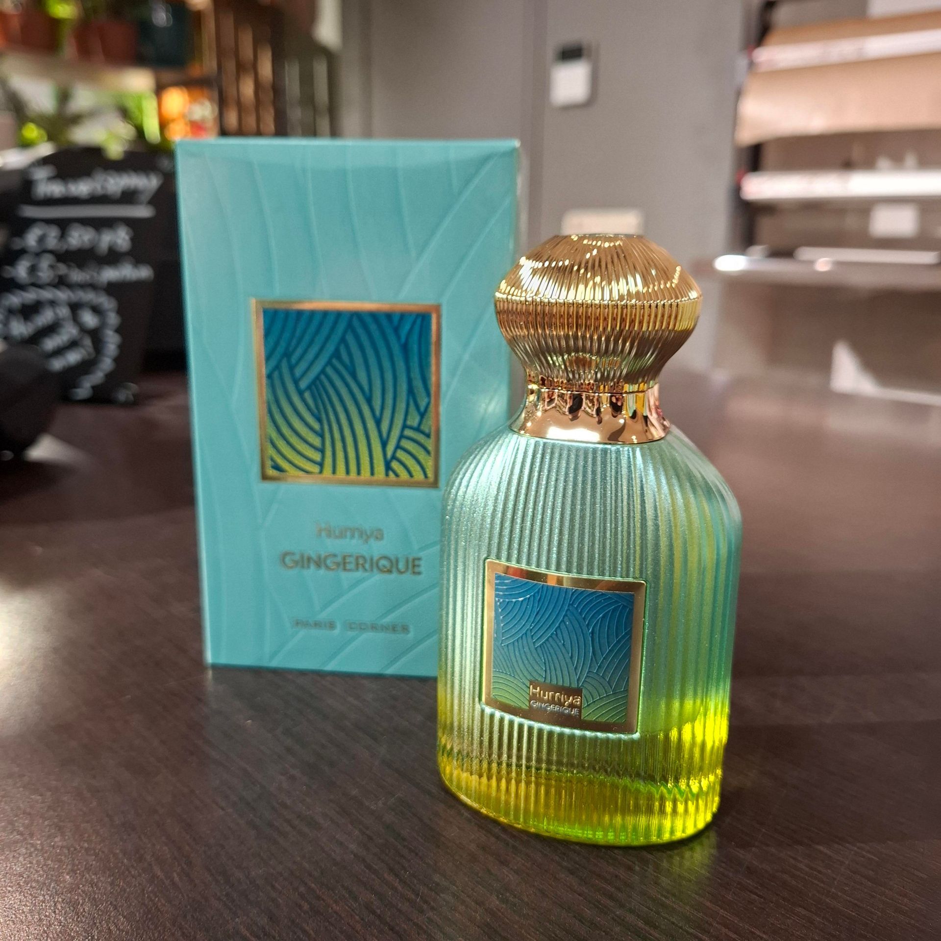 Een flesje Hamidi Gingerique parfum met een gouden dop en een glas met een kleurverloop van turquoise naar geel, naast het bijpassende doosje.