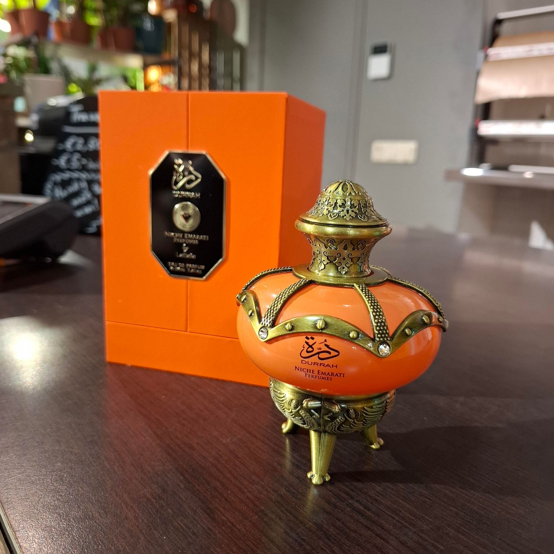 Een sierlijke, oranje parfumfles op een driepoot, naast de bijpassende oranje doos, op een donkere houten tafel.