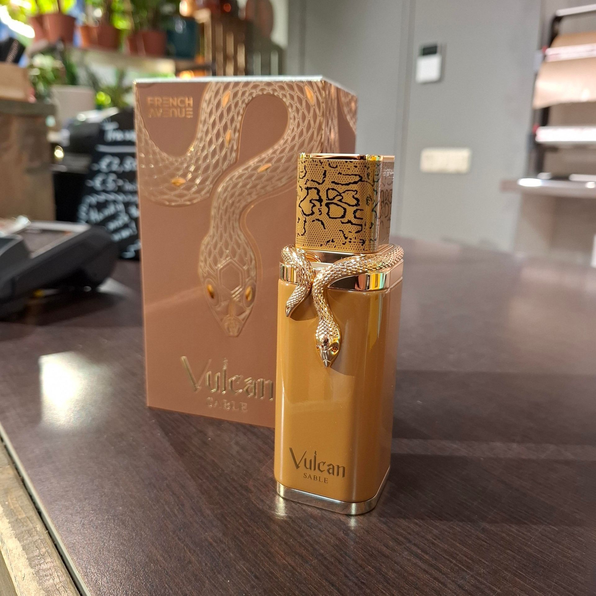 Een beige parfumflesje met een gouden slang om de hals, dat voor het bijbehorende doosje op een bureau staat.