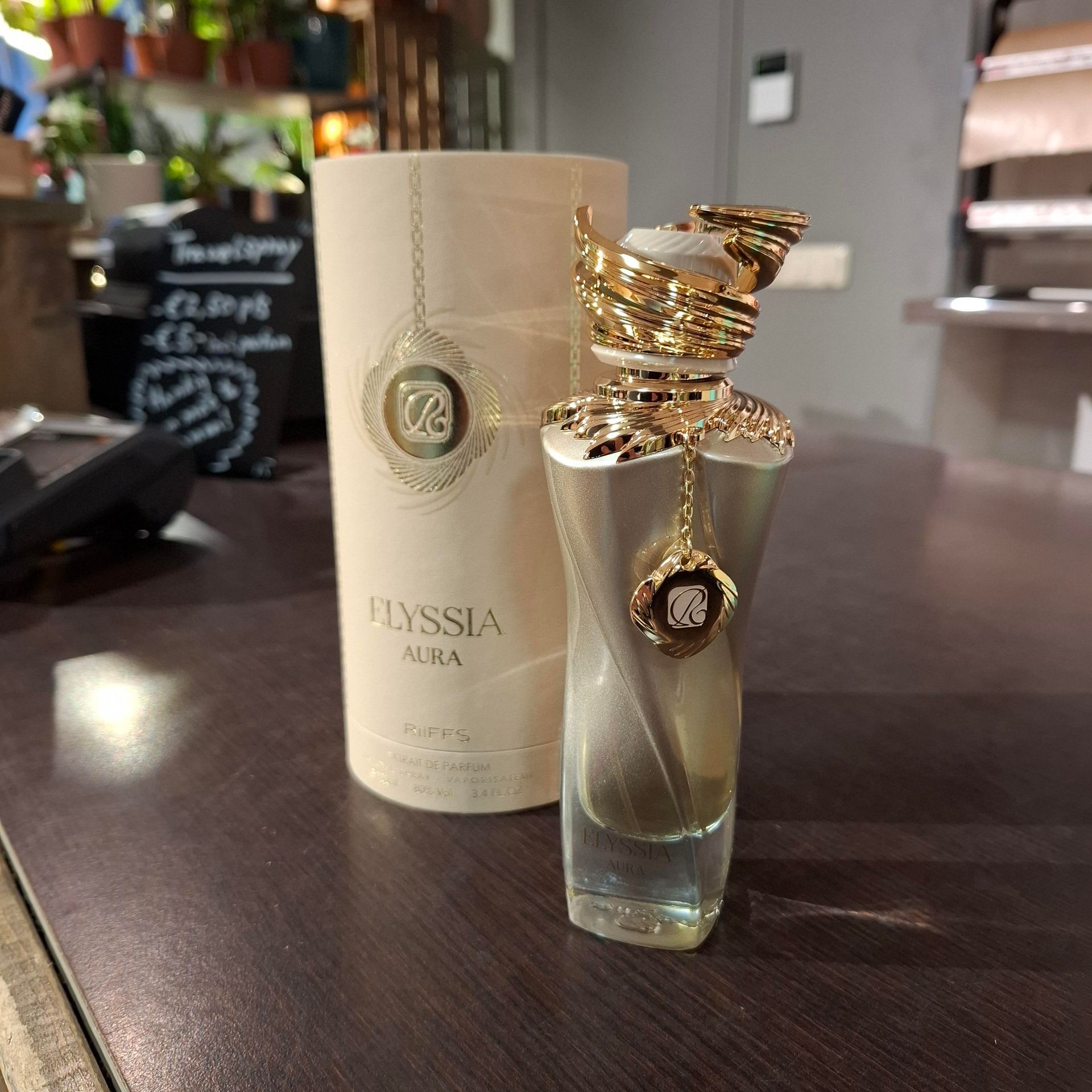 Een flesje Elyssia Aura-parfum en de bijbehorende cilindervormige doos staan ​​op een donkerhouten oppervlak in een zacht verlichte kamer.