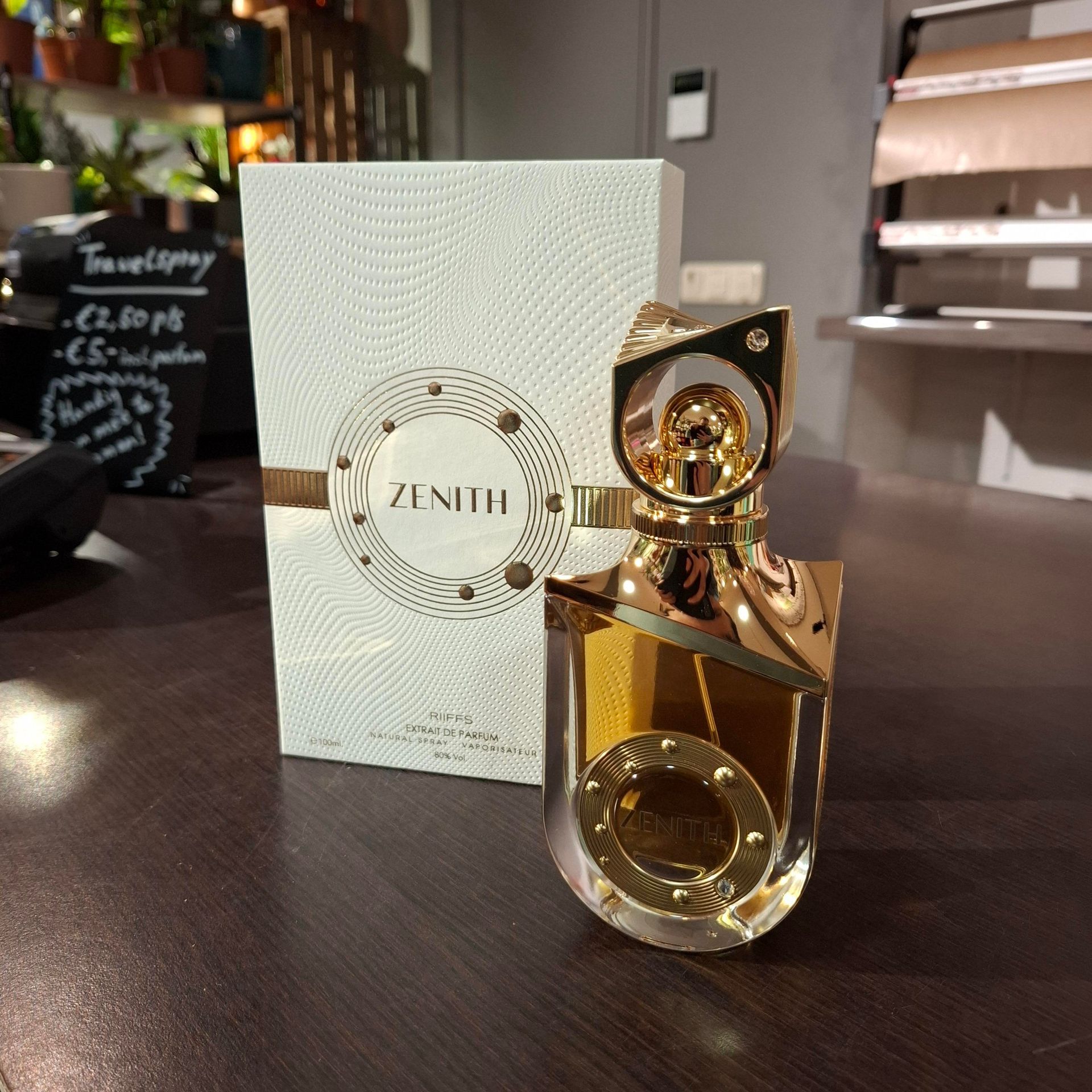 Een flesje Zenith-parfum in een goudkleurige, transparante glazen flacon staat naast de witte, gestructureerde verpakking.