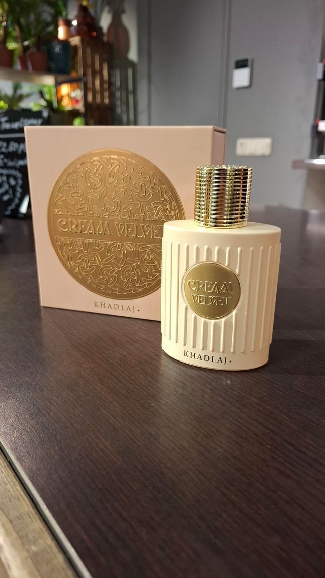 Een beige, geribbelde parfumfles met een gouden dop en een bijpassende doos met gouden etiket, staat op een donkere houten tafel.