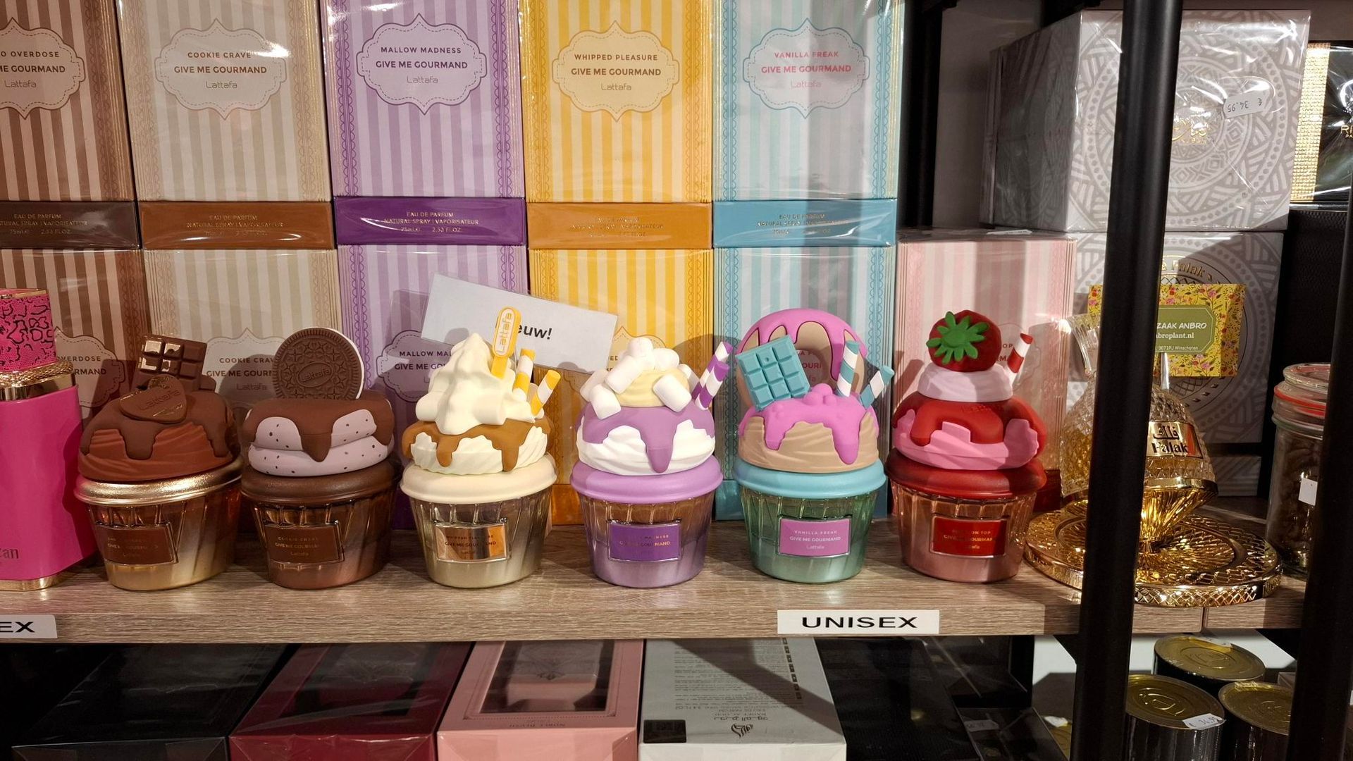 Op een plank staan ​​zes kleurrijke, cupcakevormige kaarsen in glazen potjes, die voor gestreepte productdozen zijn geplaatst.