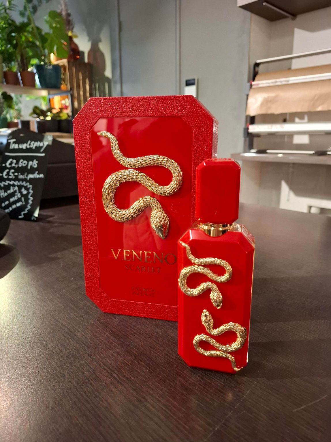 Een rode parfumfles en het bijbehorende doosje, beide versierd met sierlijke gouden slangen, staan ​​uitgestald op een houten ondergrond.