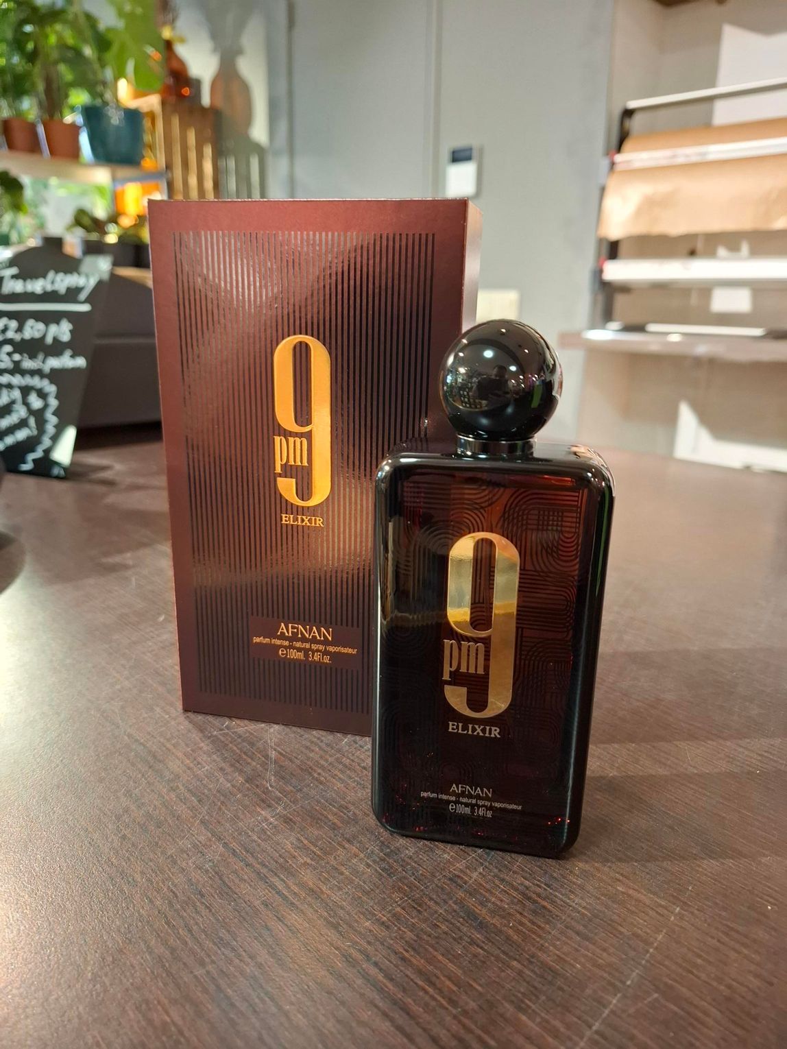 Een flesje Afnan 9 PM parfum staat naast de bijbehorende donkerrode doos op een bruine tafel.