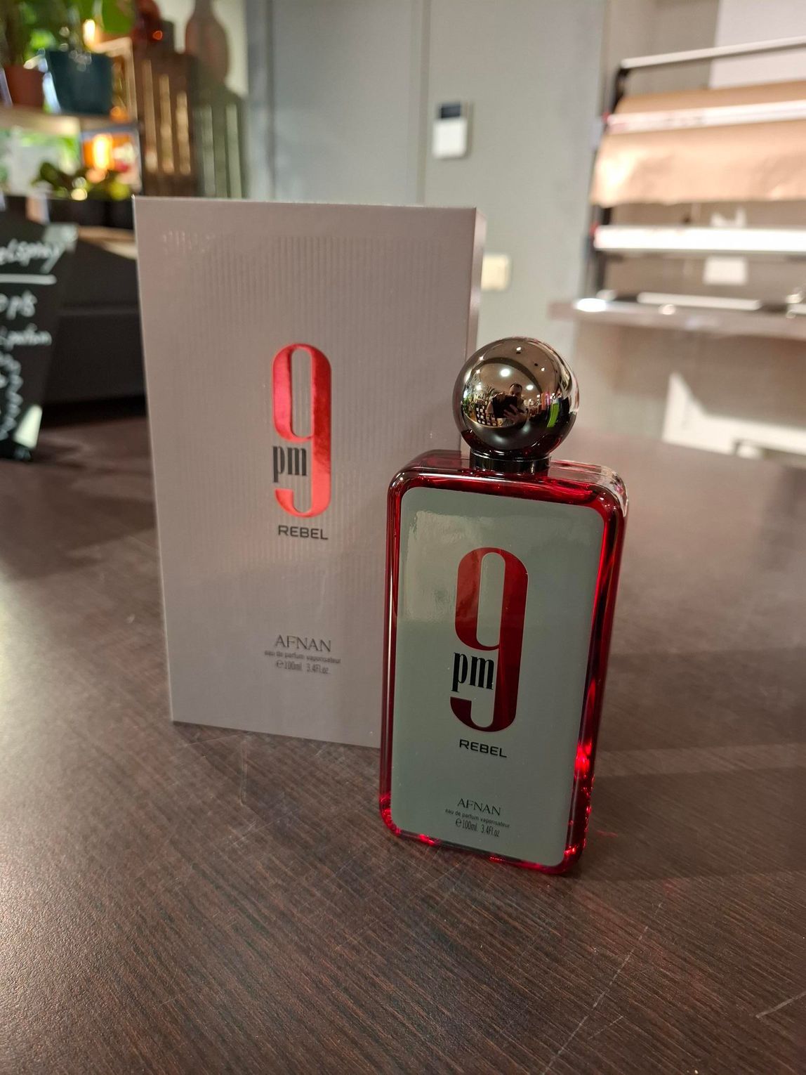 Een fles Afnan 9pm eau de cologne met de bijbehorende witte verpakking staat op een houten ondergrond in een zacht verlichte ruimte.