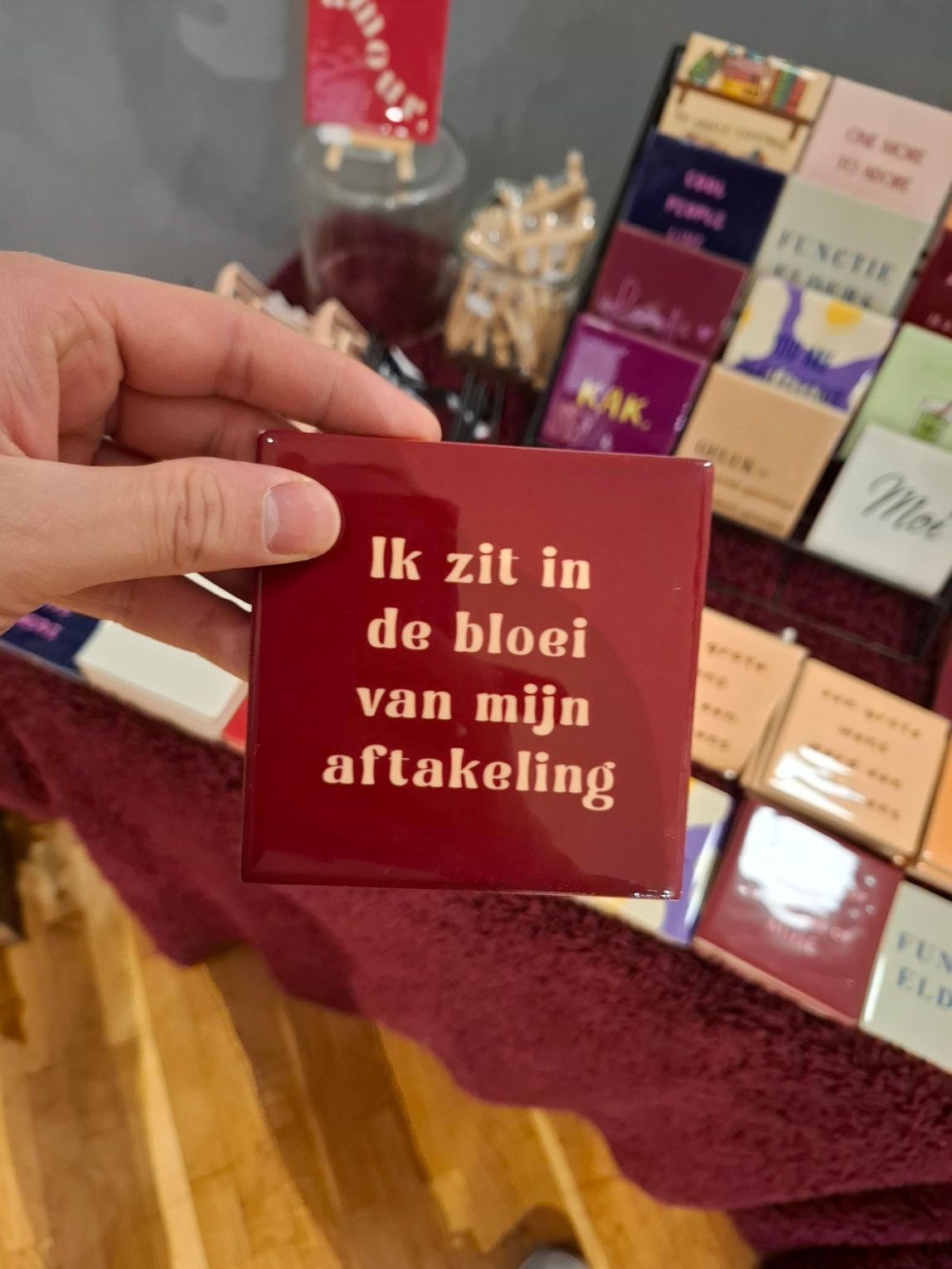 Hand holding a maroon card reading “Ik zit in de bloei van mijn afkalbing” at a gift display