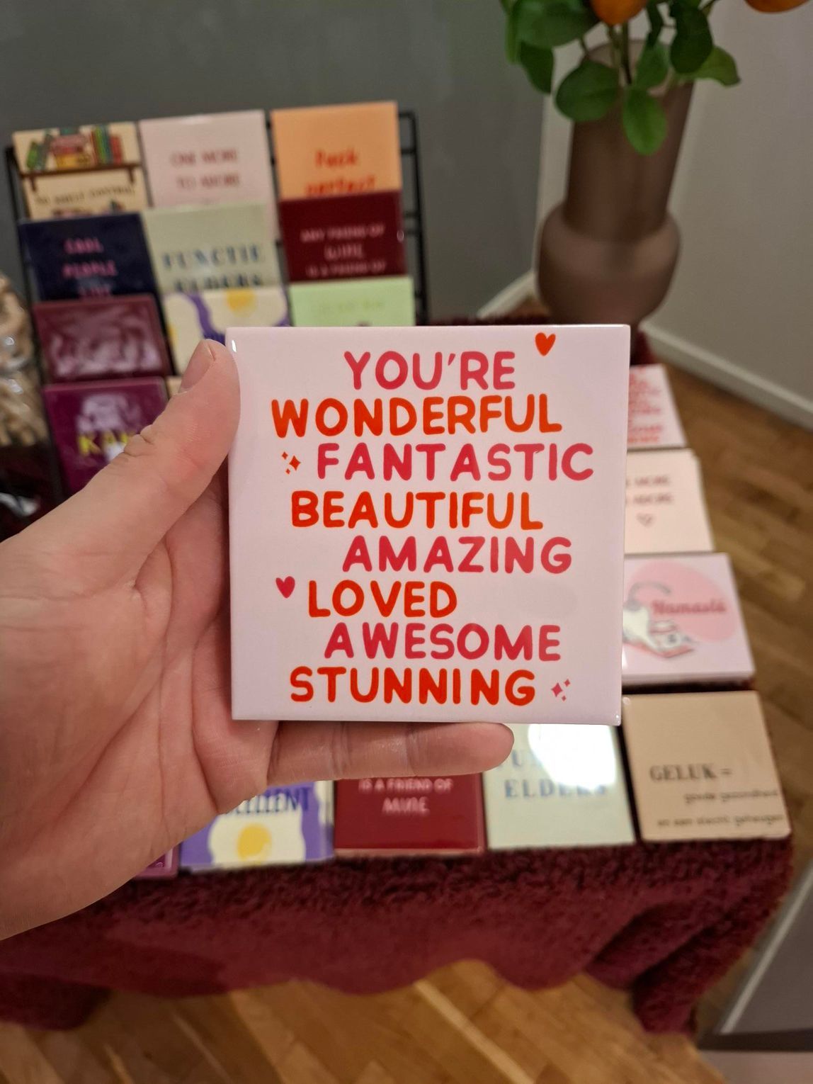 Een hand houdt een witte kaart vast met rode complimenten erop, boven netjes gerangschikte geschenkdozen en een vaas met bloemen.