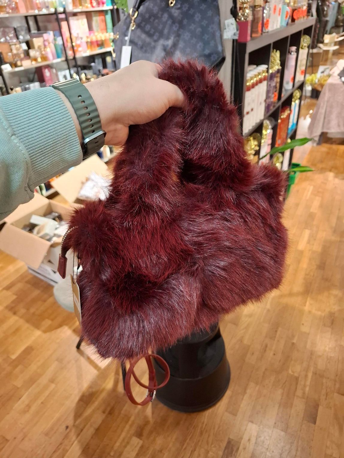 In een felverlichte winkel met schappen vol producten houdt een hand een pluizige, diep bordeauxrode handtas van imitatiebont vast.