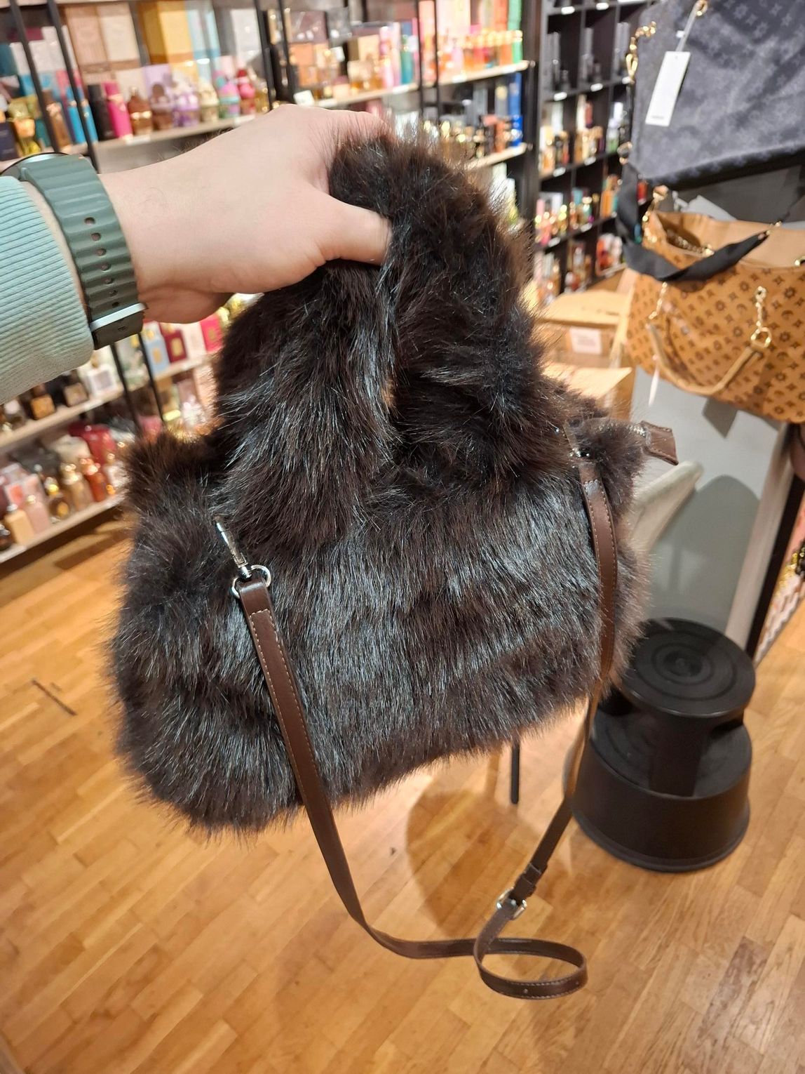 In een winkel houdt een hand een donkerbruine handtas van imitatiebont omhoog, met een dunne, bruine leren schouderband.