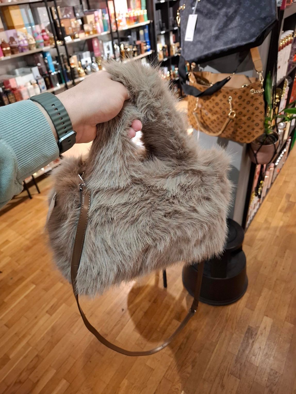 Een hand houdt een pluizige, taupekleurige handtas van imitatiebont vast met een bruine schouderband, tegen de achtergrond van een winkel.