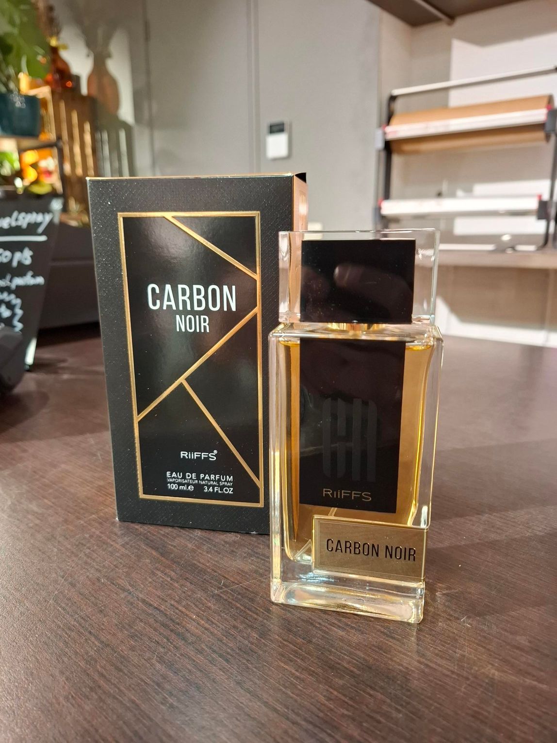 Een flesje parfum Carbon Noir naast de zwart-gouden doos op een houten tafel.