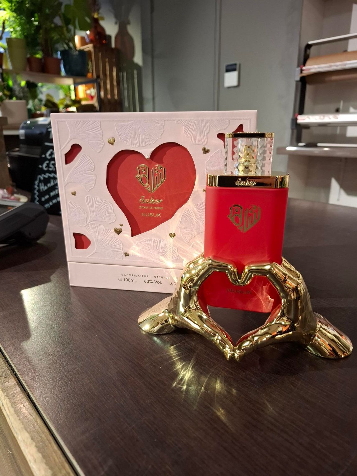 Een rode parfumfles staat in een gouden, hartvormige houder, met op de achtergrond een bijpassende wit-rode geschenkdoos.