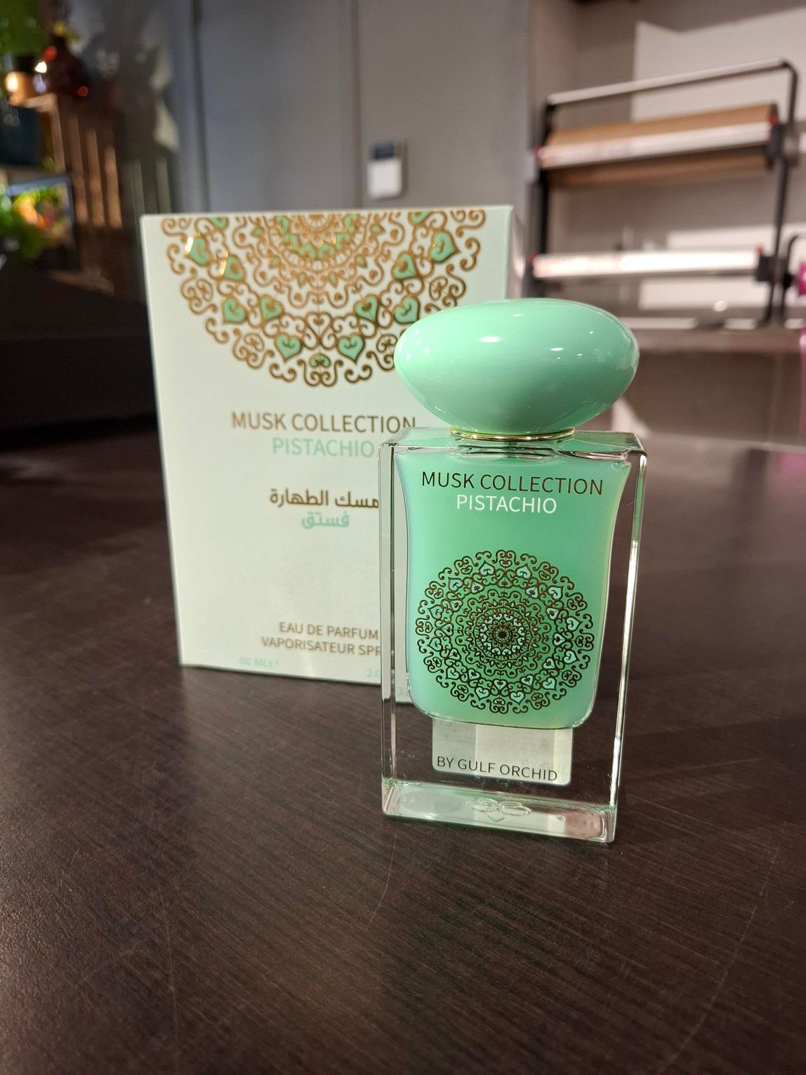 Een flesje parfum Musk Collection Pistachio staat naast de bijpassende wit-gouden doos met patroon op een tafel.