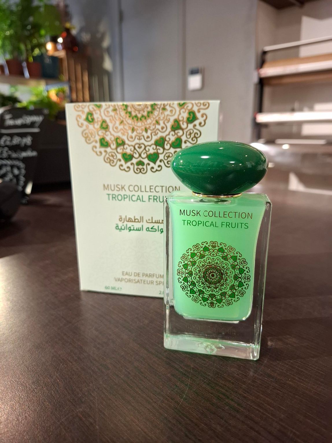 Een flesje parfum Musk Collection Tropical Fruits staat voor de witte doos op een donkerhouten ondergrond.