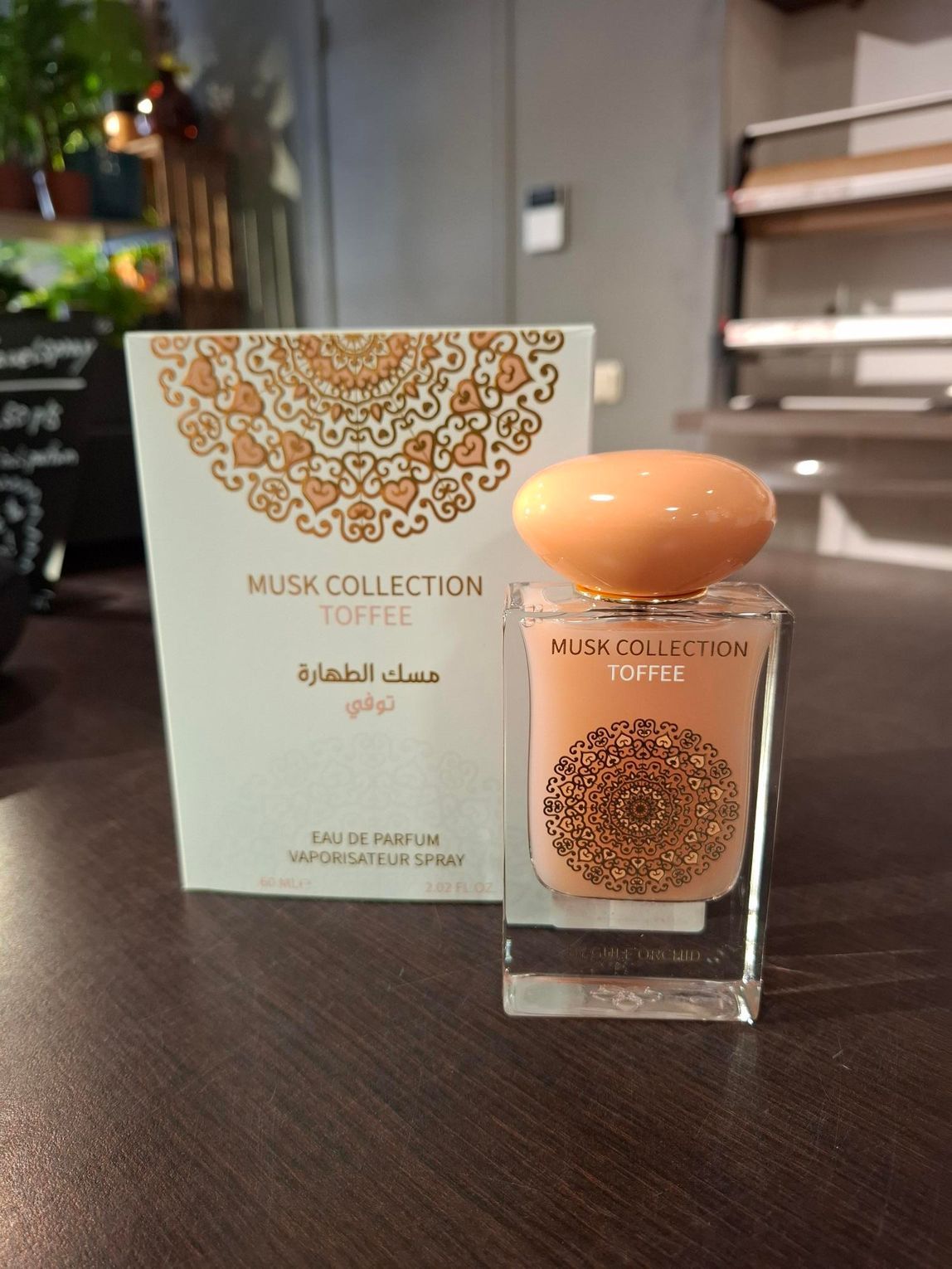 Een flesje parfum Musk Collection Toffee en de bijbehorende decoratieve witte doos staan ​​op een houten tafel in een schemerig verlichte ruimte.
