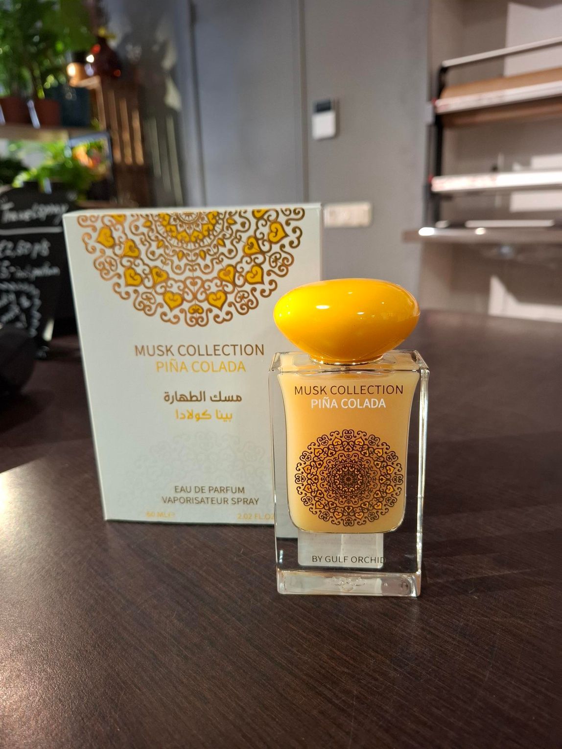 Een flesje Pina Colada-parfum van de Musk Collection en de bijbehorende witte doos met gouden mandala-motieven op een donkere tafel.