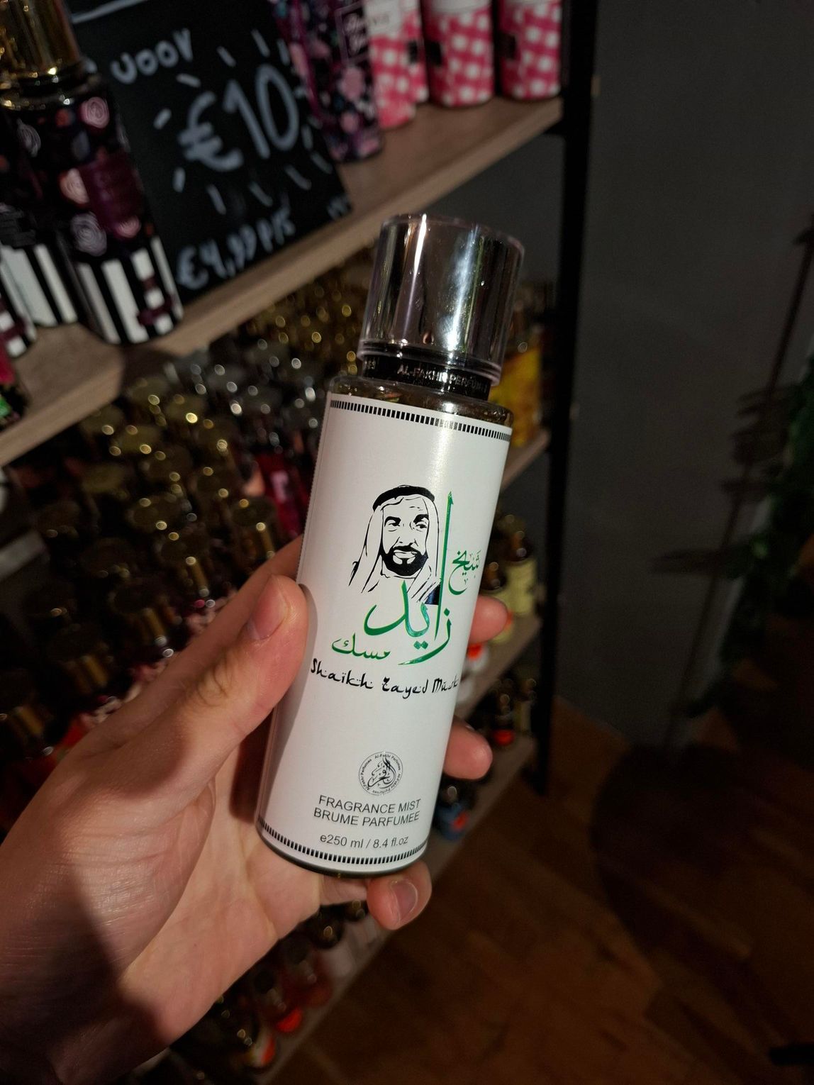 In een winkel houdt iemand een flesje parfum van Zayed Musk vast, met daarop een afbeelding van een man en groene Arabische kalligrafie.