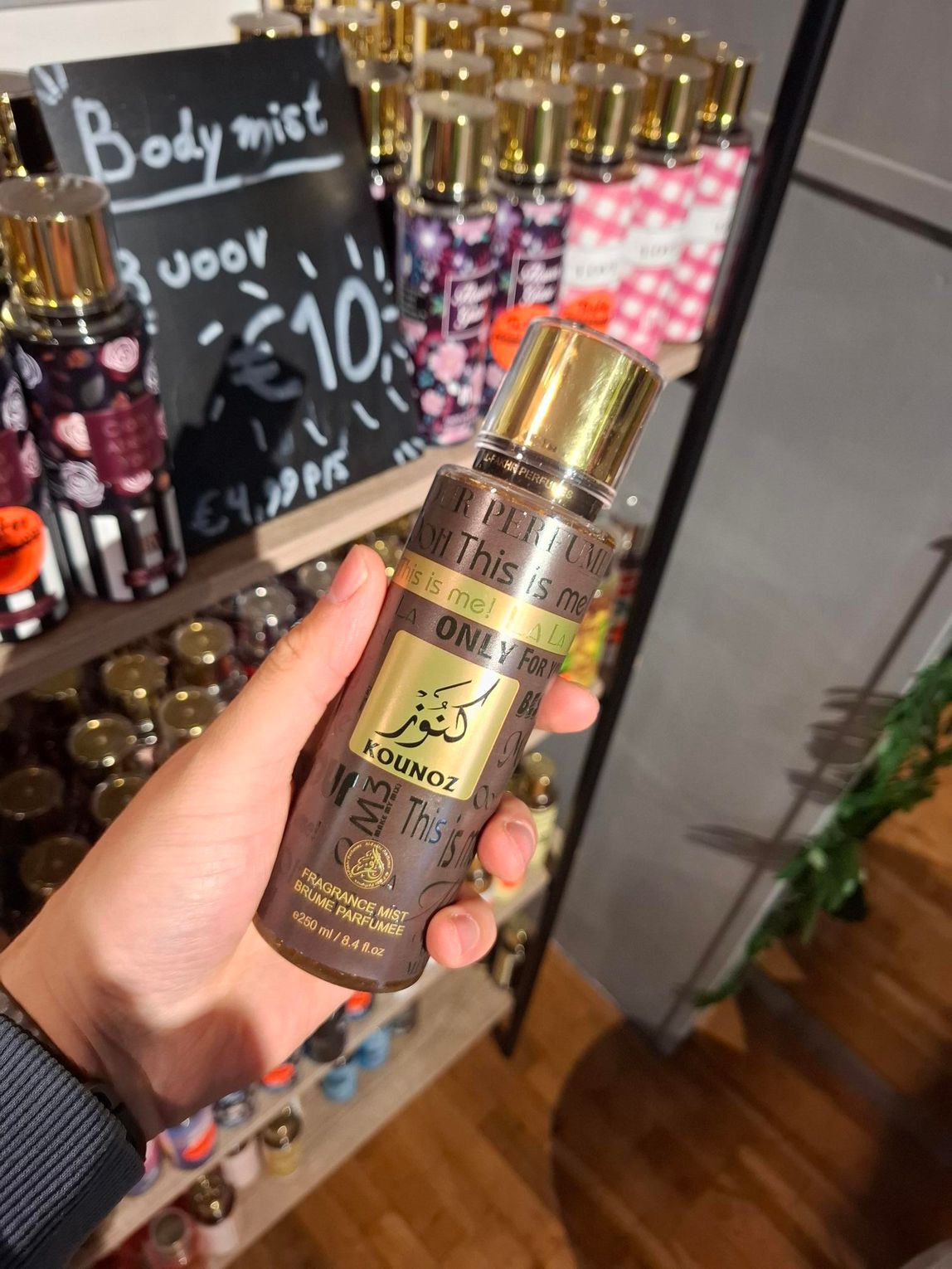 In een winkel houdt iemand een bruine fles Kanz Al Oud body mist vast, met op de achtergrond schappen vol body mists.