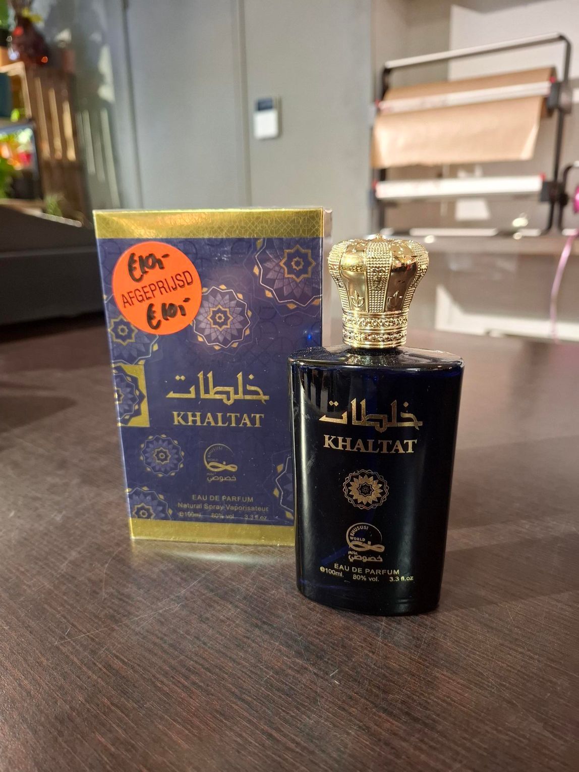 Een zwart flesje KHAFAJAT-parfum met gouden dop, naast een bijpassende doos, staat op een tafel.
