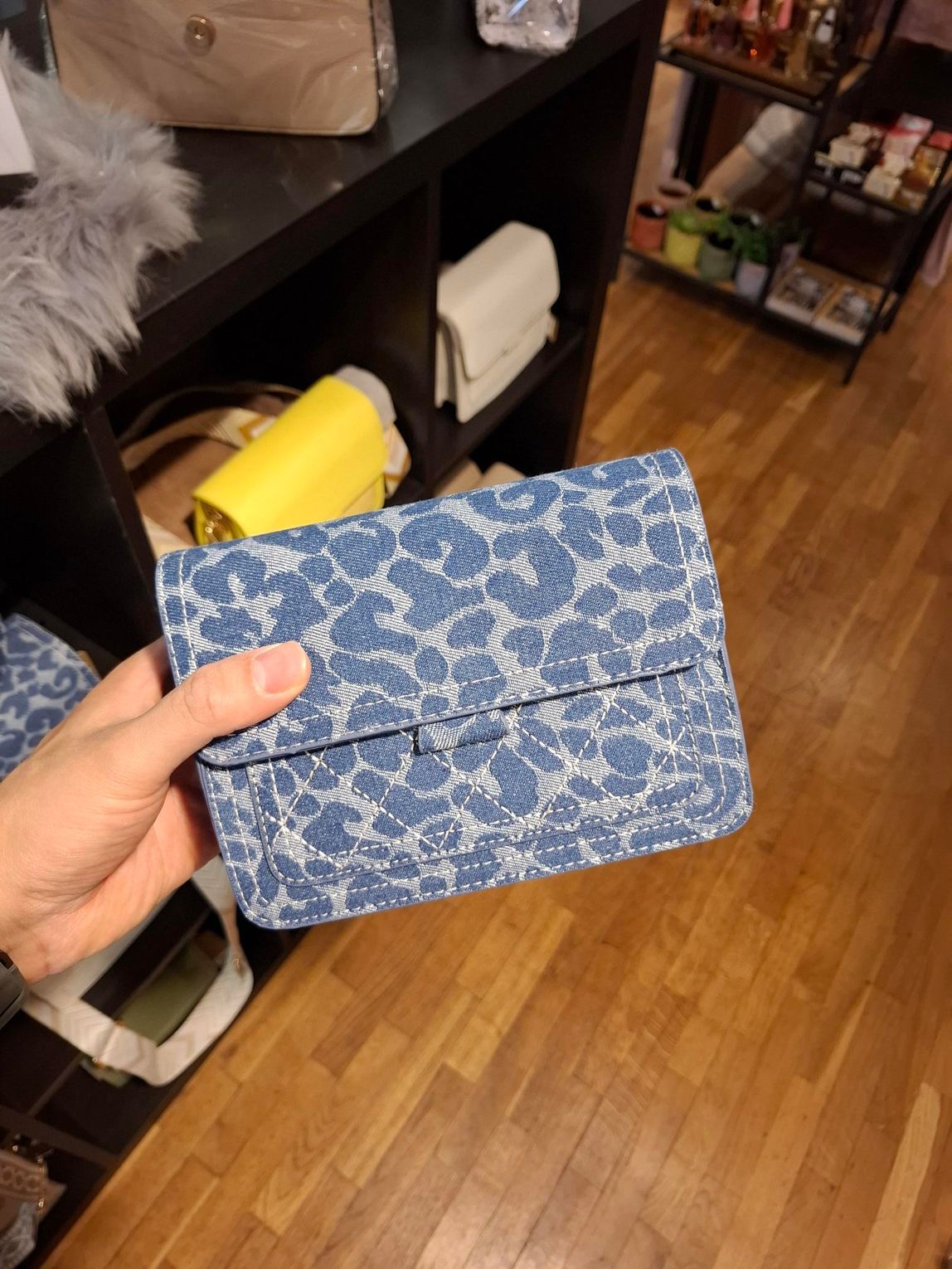 Hand die een blauwe portemonnee met patroon vasthoudt boven een houten vloer in een winkel of interieur van een woning.